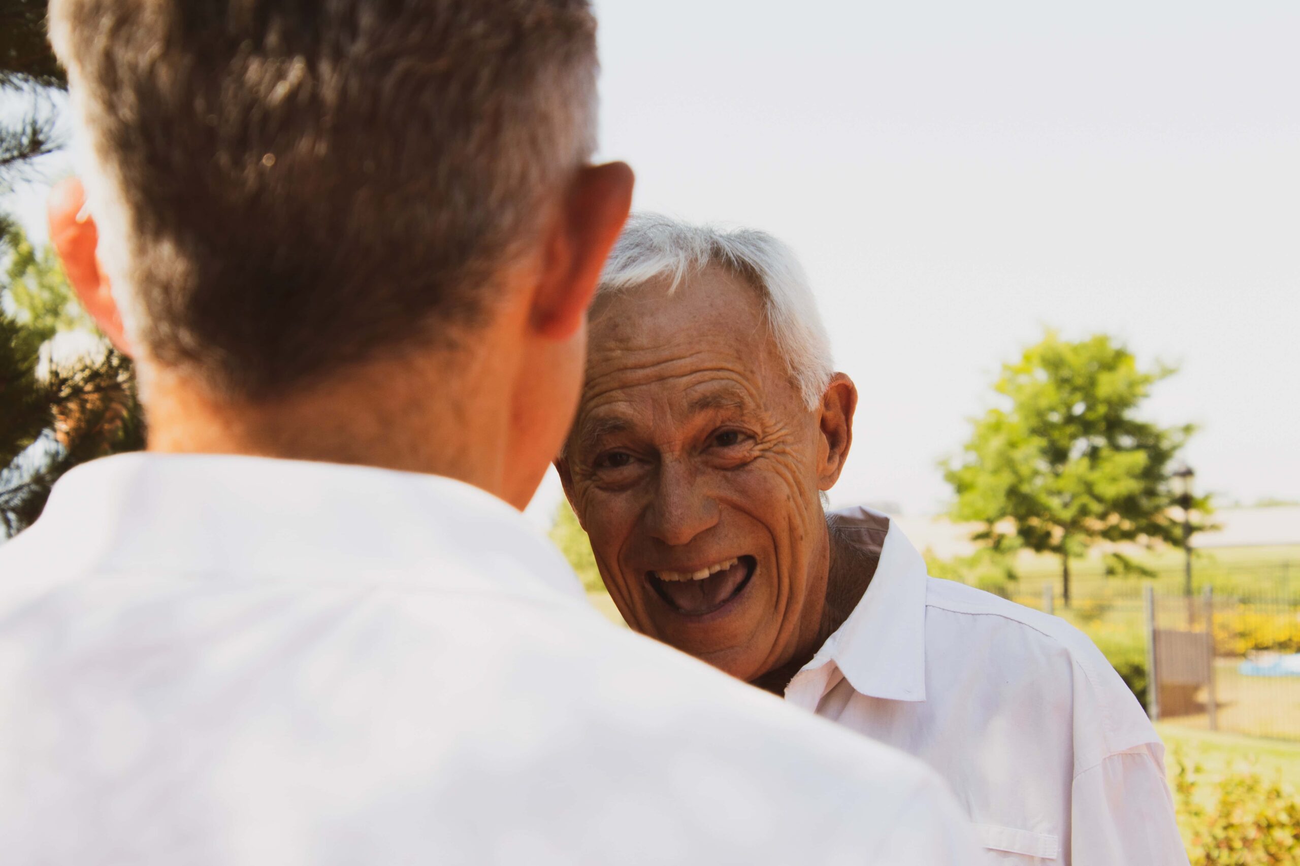 Two older men in white shirts talking outdoors; one faces the camera smiling, while the other’s back is visible. Trees and a fence are in the background.