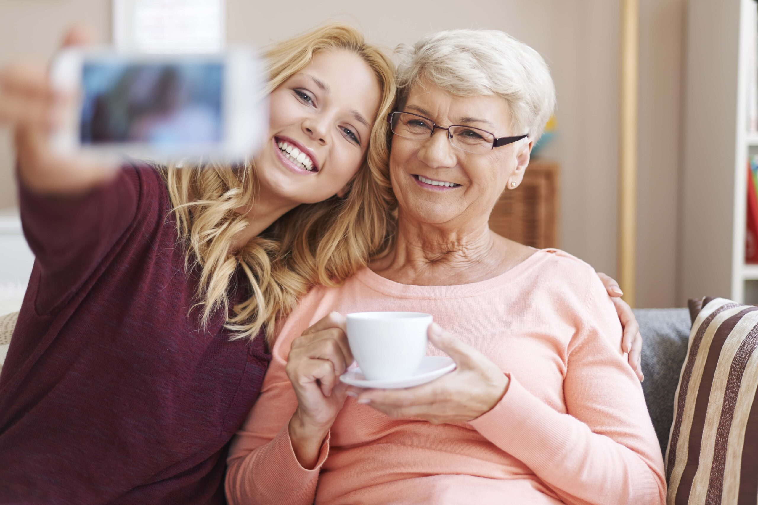 A young woman taking a selfie with an older woman who is holding a cup, both smiling and sitting together on a couch.