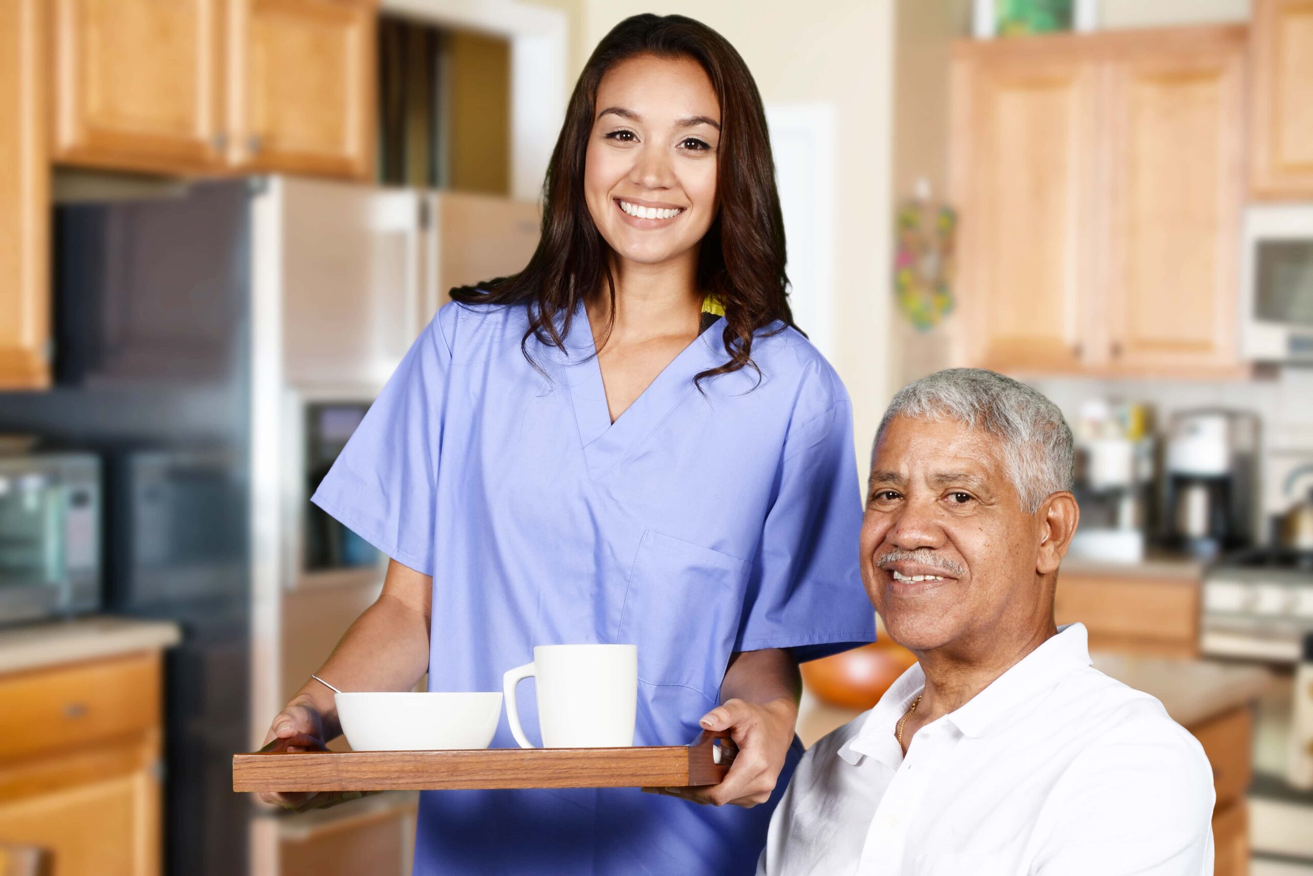 A caregiver in scrubs holds a tray with a bowl and mug, standing beside an older man seated in a kitchen. Both are smiling at the camera.