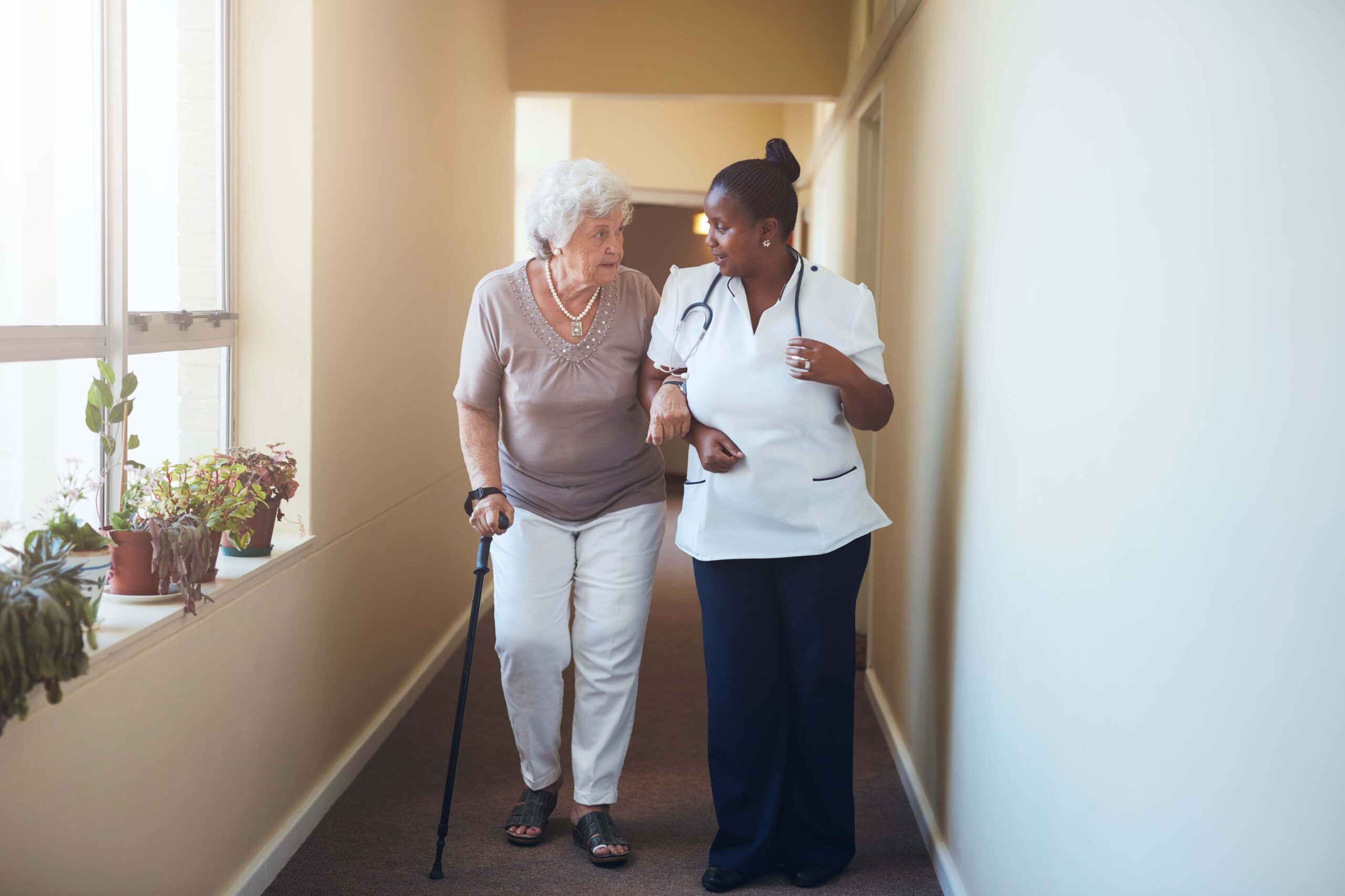 An elderly woman with a cane walks down a hallway, assisted by a healthcare worker in uniform.