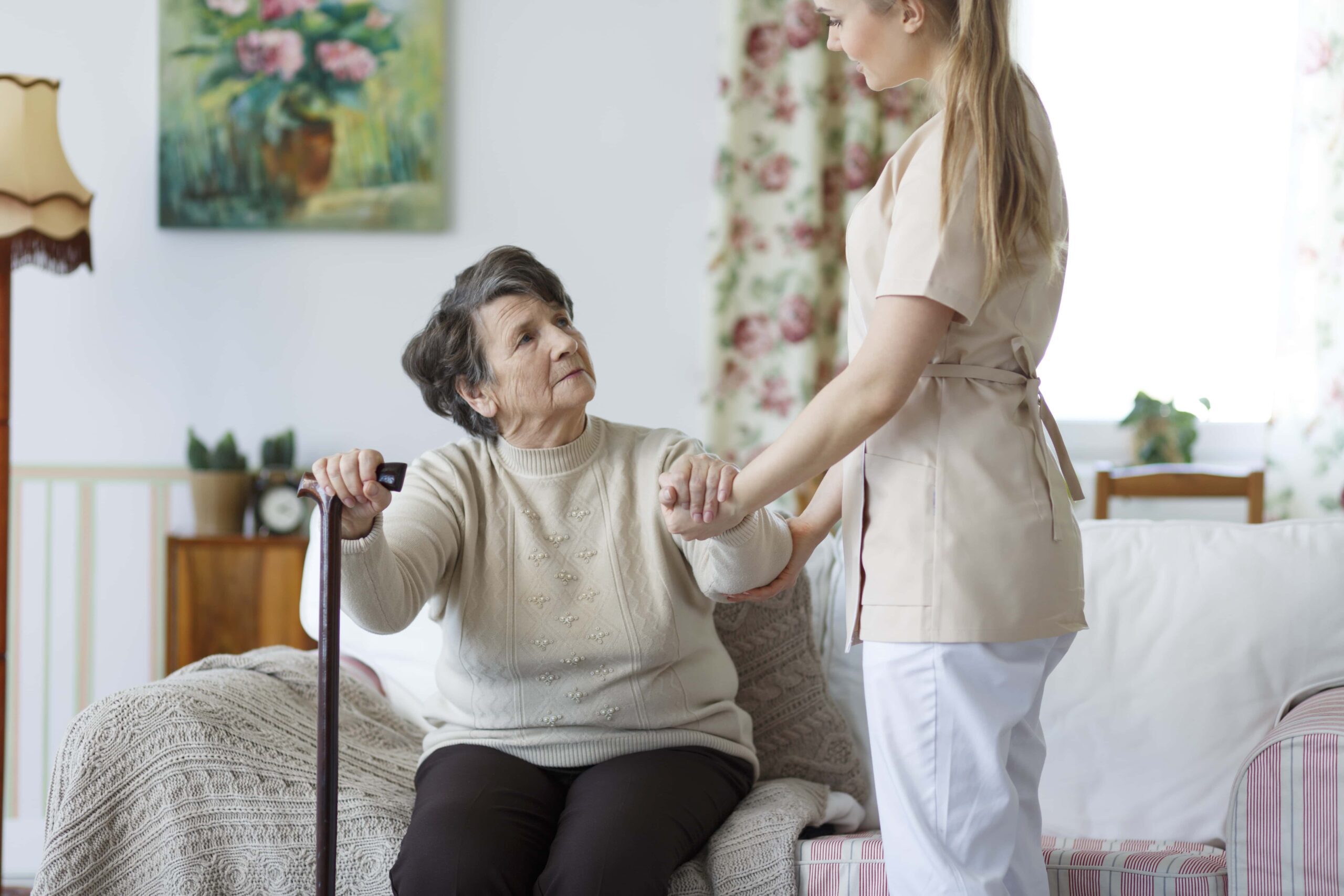 An elderly woman with a cane sits on a couch and holds hands with a caregiver standing beside her in a home setting.