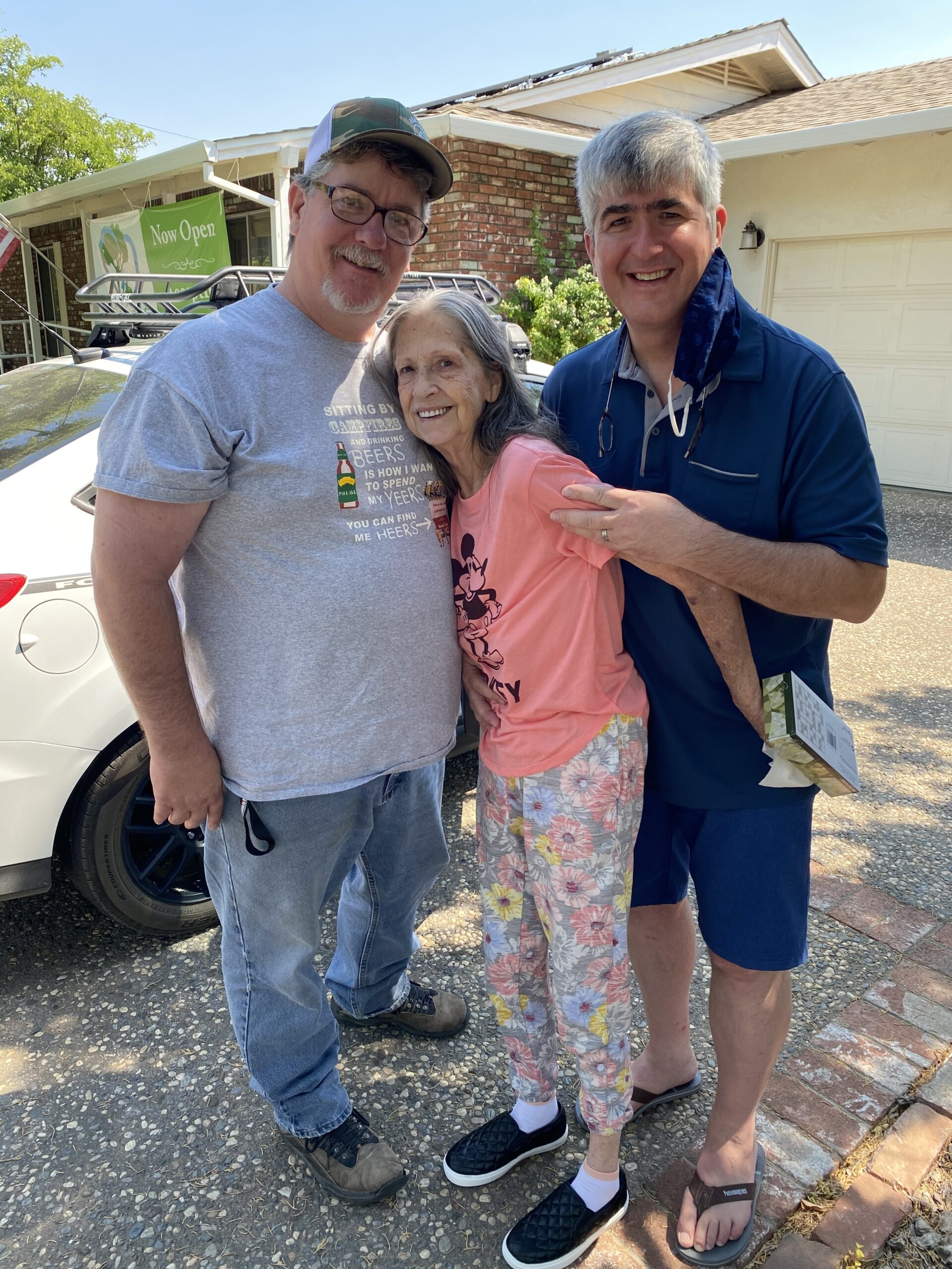 Three people stand together outside near a white car and a house, smiling at the camera on a sunny day.