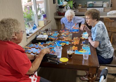 Three elderly women sit around a table working on a jigsaw puzzle in a kitchen, with drinks and walkers nearby.
