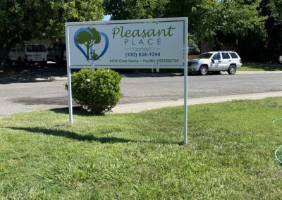 A sign on a grassy lawn reads "Pleasant Place," displaying contact information and license details, with trees, cars, and a street in the background.