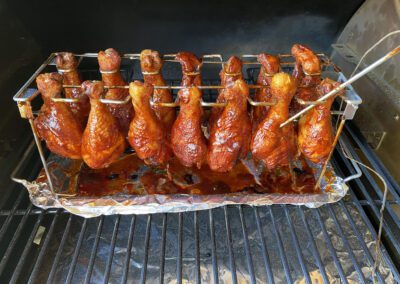 Chicken drumsticks coated in sauce are arranged vertically on a metal rack inside a grill, with a meat thermometer inserted into one drumstick.