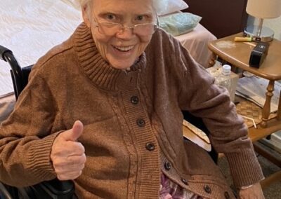 Elderly woman with white hair and glasses sits in a wheelchair, wearing a brown sweater, smiling, and giving a thumbs up in a cozy room.