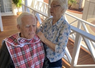 An elderly woman stands next to an elderly man in a wheelchair, fastening a red plaid bib around his neck on a wooden porch.