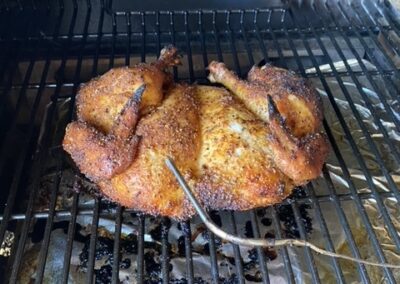A seasoned spatchcock chicken is cooking on a grill with a meat thermometer probe inserted, surrounded by metal grates.