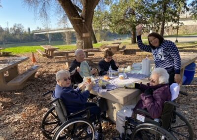 Five people, including two in wheelchairs, sit at a picnic table outdoors, eating lunch. One person stands and waves at the camera. Trees and a bridge are visible in the background.