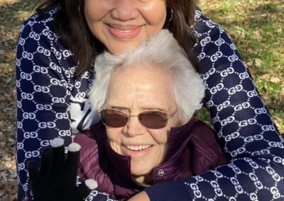 Two women are outdoors on a sunny day. The younger woman stands behind and hugs the older woman, who is wearing sunglasses and a purple jacket. They are both smiling.