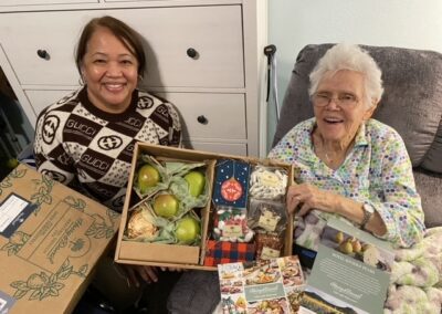 Two women sit together, smiling, with an open gift box containing fruit, snacks, and brochures on their laps in a cozy indoor setting.