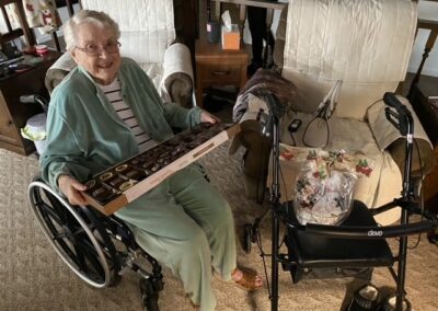 An elderly woman in a wheelchair holds an open box of assorted chocolates, seated next to a walker and a wrapped gift in a living room.