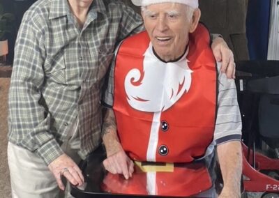 An elderly woman stands next to an elderly man in a wheelchair. Both wear Santa hats, and the man has a festive Santa-themed bib. They are smiling indoors.