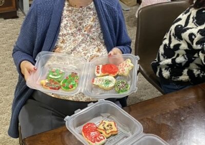 An elderly woman sits at a table holding two containers of decorated Christmas cookies, with more cookies in a container on the table in front of her.