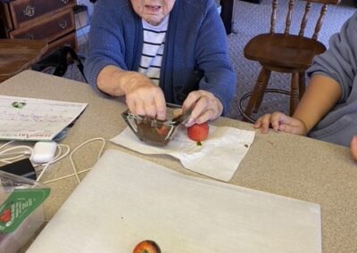 An older woman dips strawberries in chocolate at a kitchen counter, while another person sits beside her. Chocolate-covered strawberries are on a tray in the foreground.