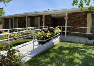 A brick house with a wheelchair-accessible ramp, white railings, flower planters along the ramp, and green grass in the yard.
