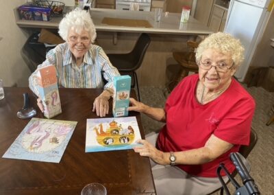 Two elderly women sit at a table assembling a Noah's Ark puzzle, with puzzle boxes and a completed US map puzzle also visible.