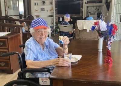 Elderly woman in a wheelchair wearing a patriotic hat sits at a table with food, holding a cupcake in a decorated room with red, white, and blue flowers.