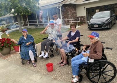 Five elderly people, three in wheelchairs, sit outside a house holding sparklers; some wear patriotic hats and an American flag is visible in the background.