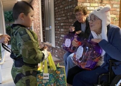 A child in a camouflage costume hands a treat bag to an elderly woman in a wheelchair wearing a white animal hat, while another woman sits nearby.