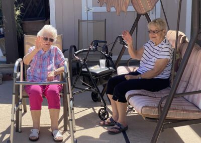 Two elderly women sit outside, one in a chair with a walker and the other on a swing, both smiling and waving at the camera. A walker and a small table are nearby.