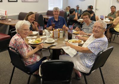 Five people sit around a round table in a restaurant, eating meals and smiling at the camera. Other diners are visible in the background.