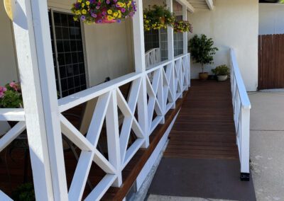 A wooden porch with white railing features hanging flower baskets and a ramp leading up to the entrance of a white house.