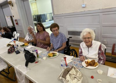 Four women sit at a long table with plates of food and bingo cards in a community hall.