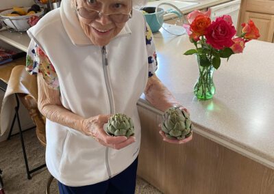 An elderly woman stands in a kitchen holding two artichokes, smiling at the camera. A vase of roses sits on the countertop behind her.