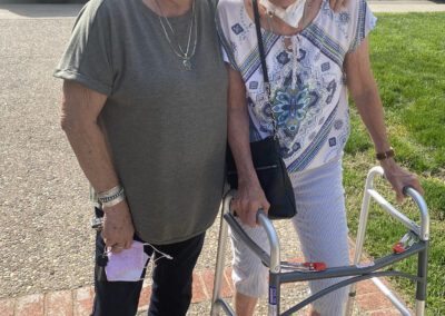 Two older women stand outside smiling, one using a walker and the other holding a face mask. They appear to be enjoying a sunny day together in a residential neighborhood.