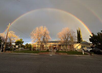 A double rainbow arches over a single-story house with leafless trees in the front yard on a suburban street.