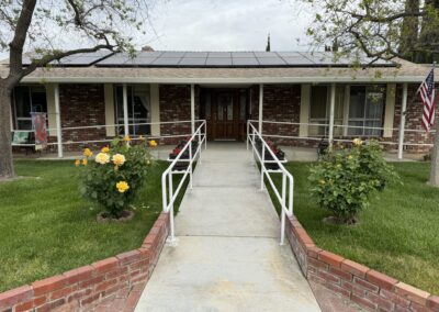 A brick house with a wheelchair-accessible ramp, solar panels on the roof, blooming yellow roses, and an American flag displayed near the entrance.