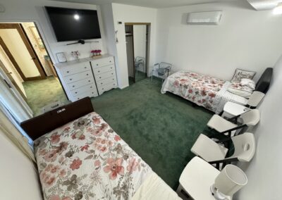A bedroom with two single beds, floral bedding, green carpet, a TV mounted above white dressers, chairs lined along one wall, and a small table between the beds.