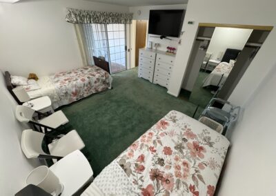 A bedroom with two single beds, floral bedding, green carpet, white dresser, TV, chairs, a walker, and a large mirror; sliding glass doors lead outside.