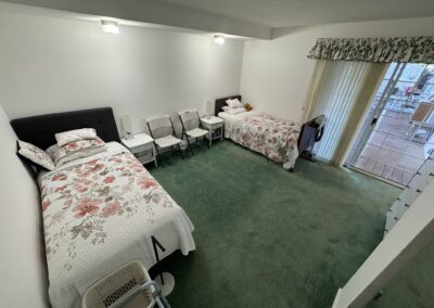 A bedroom with two single beds with floral bedding, several chairs between them, green carpet, and sliding glass doors leading to a deck outside.