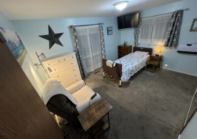 A tidy bedroom with a hospital bed, wooden furniture, a recliner, dresser, lamp, TV mounted on the wall, and two windows with curtains on light blue walls.