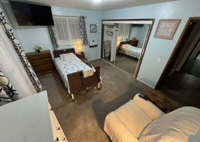 A small, tidy bedroom with a hospital bed, recliner, mirrored closet, dresser, TV, and medical equipment on a carpeted floor.