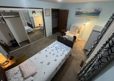 A small bedroom with a single bed, mirrored closet doors, a dresser, an armchair with a lamp, and a beach landscape painting on the wall.