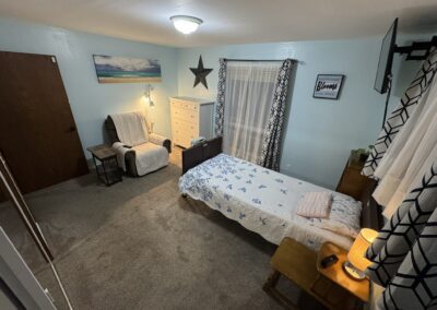 A neatly arranged bedroom with a single bed, armchair, dresser, and window with sheer curtains; a lamp, wall art, and a TV are also visible.