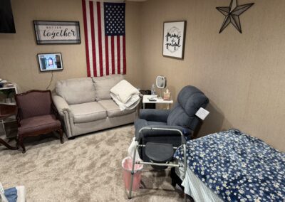 A cozy room with a beige couch, recliner, hospital bed, walker, American flag, and wall decor. A TV and framed sign are also visible. Carpeted floor and neutral walls.