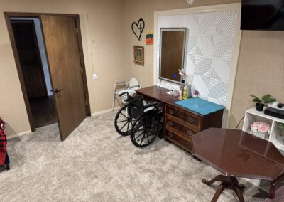 A wheelchair is positioned in front of a dresser with a mirror in a carpeted room; a walker is near an open door, and a table is in the foreground.