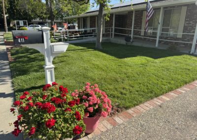 A white mailbox labeled 411 stands near red flowers in pots, in front of a single-story brick house with a green lawn and an American flag.