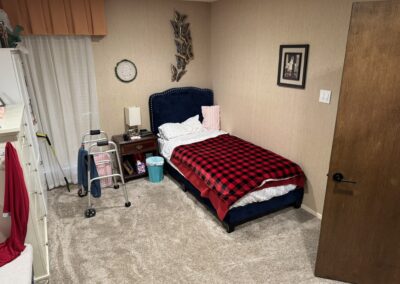 A small bedroom with a single bed covered by a red and black plaid blanket, a nightstand, walker, dresser, wall clock, framed picture, and beige carpet.