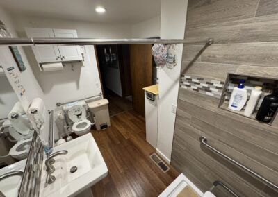 A bathroom with a sink, toilet, child potty seat, grab bars, wooden floor, wall shelf with toiletries, and white cabinets.