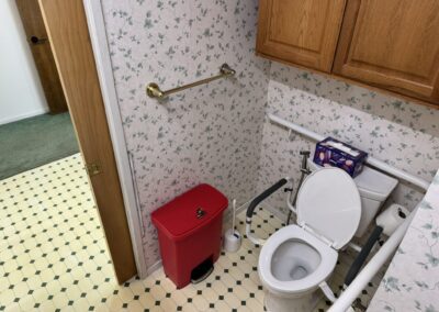 A small bathroom with floral wallpaper, an open toilet with support rails, a red trash bin, a roll of toilet paper, tissues, and wooden cabinets above.