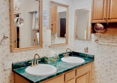 Bathroom with dual sinks, green countertop, three mirrors, wooden cabinets, floral wallpaper, and patterned tile floor. Ceiling lights and paper towel holder are visible.
