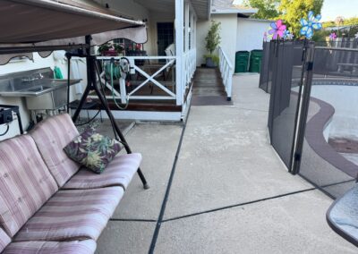Outdoor patio with a striped swing bench, a pillow, outdoor sink, ramp to the house, three green bins, and a fenced-off empty pool area with colorful pinwheel decorations.