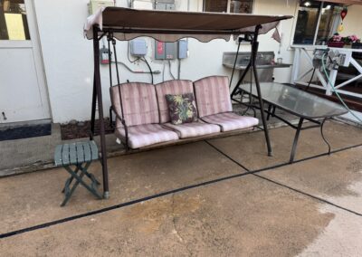 Outdoor patio swing with three striped cushions and a floral pillow, next to a glass-topped table and a small green side table, positioned on a concrete surface by a house wall.