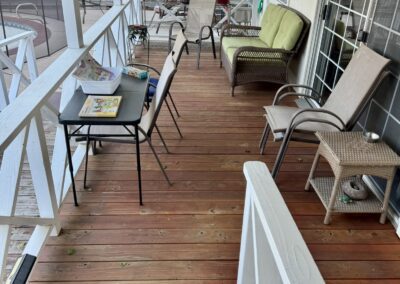 A wooden porch with chairs, a small table with papers, a loveseat, potted plants, and a view of a shed and backyard in the background.
