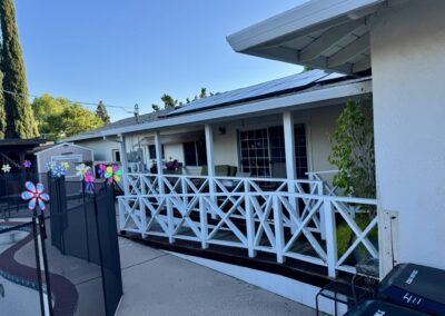 A white house with solar panels on the roof, a fenced porch, and outdoor seating. A pool and safety fence are in the foreground, with trees and a shed in the background.